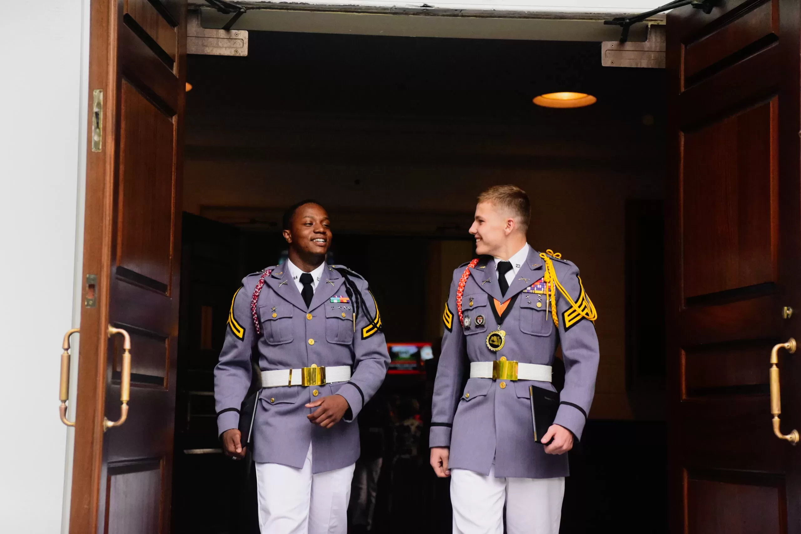 Two cadets in dress uniform walking side by side through a doorway.